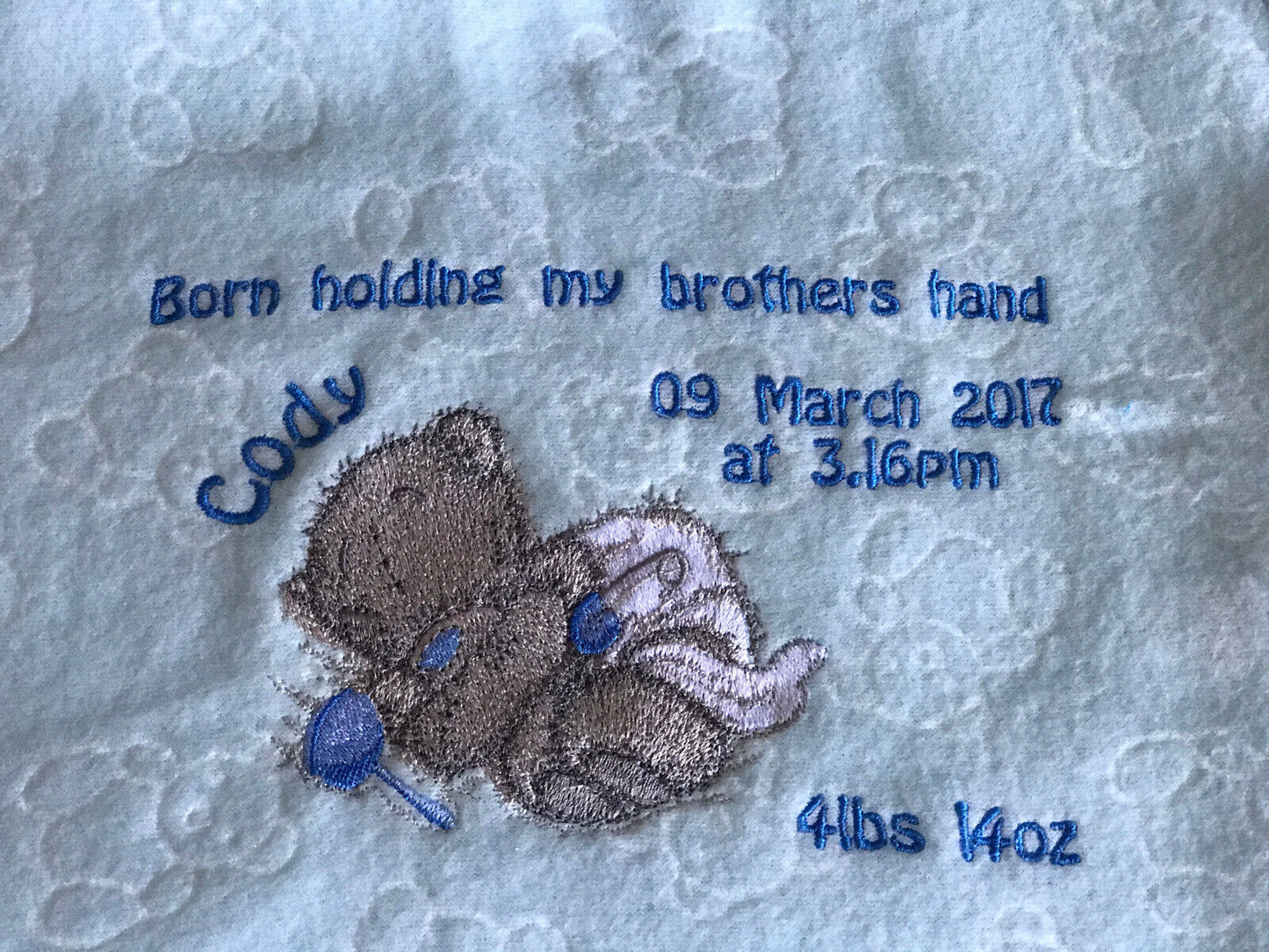 Grey Tatty Teddy asleep in a nappy, embroidered on a light blue fleece blanket with a blue rattle.