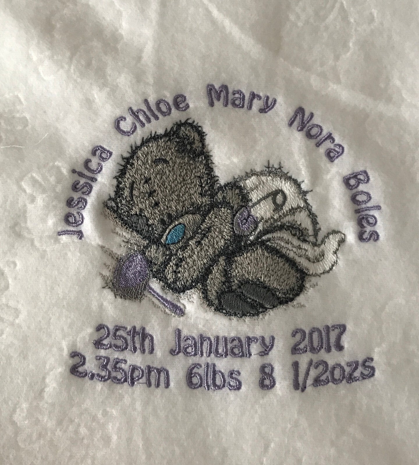Grey Tatty Teddy asleep in a nappy, embroidered on a white embossed fleece blanket with a lilac rattle.