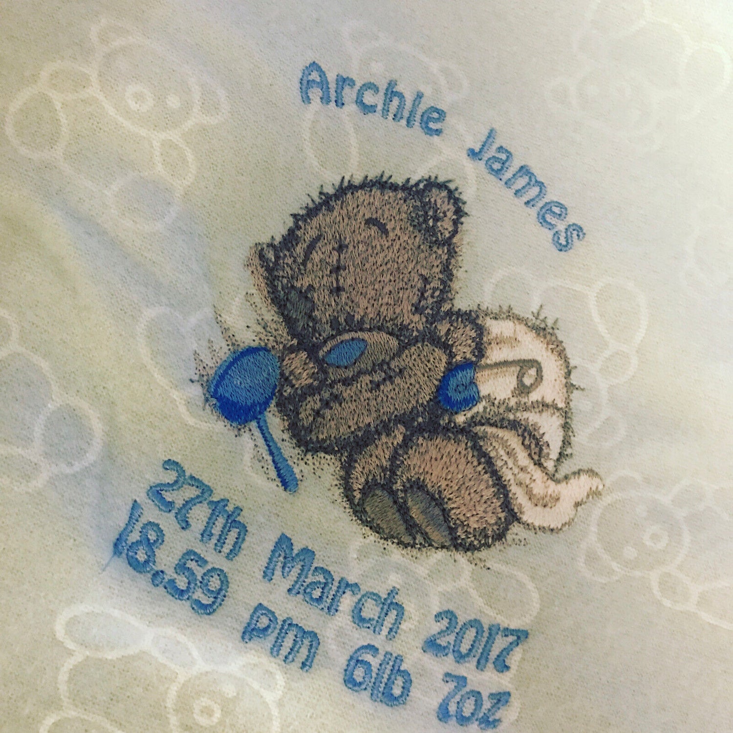 Grey Tatty Teddy asleep in a nappy, embroidered on a light blue fleece blanket with a blue rattle.